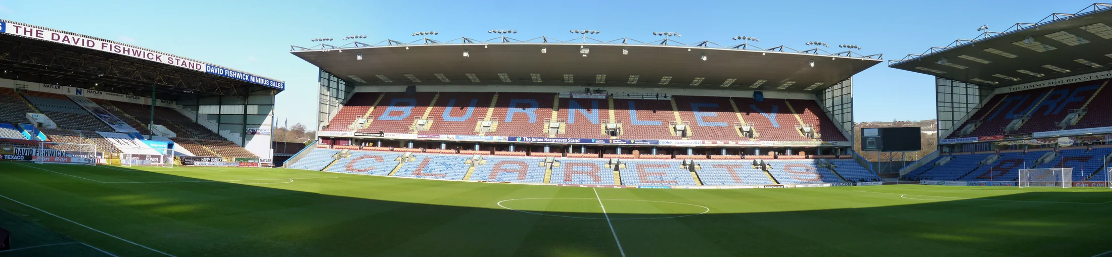 Wide interior panorama of Turf Moor, with BURNLEY CLARETS picked out across the claret and blue seats.